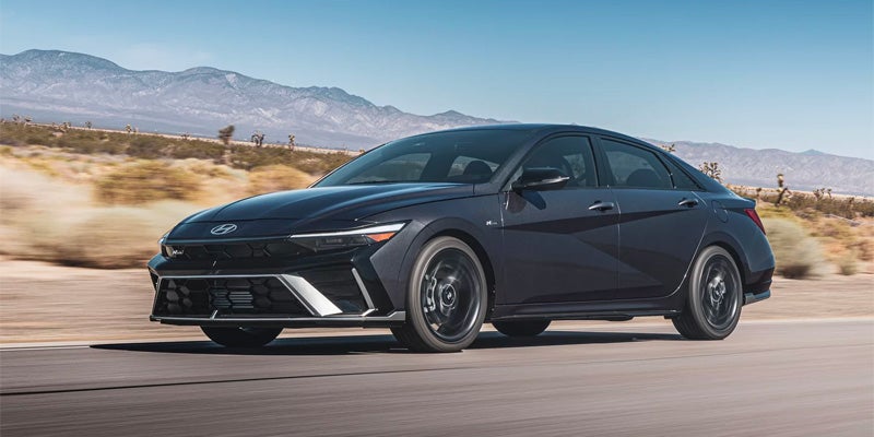 A dark gray Hyundai Elantra drives right on a paved road with blurred desert landscape and mountains in the background
