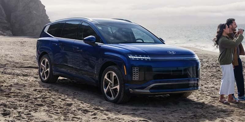 A navy blue Hyundai Ioniq 7 parked on a sandy beach, with a couple looking at the ocean in the background