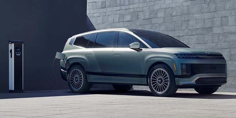 A teal electric SUV charging from a white and black station on a grey concrete floor, against a concrete-block wall