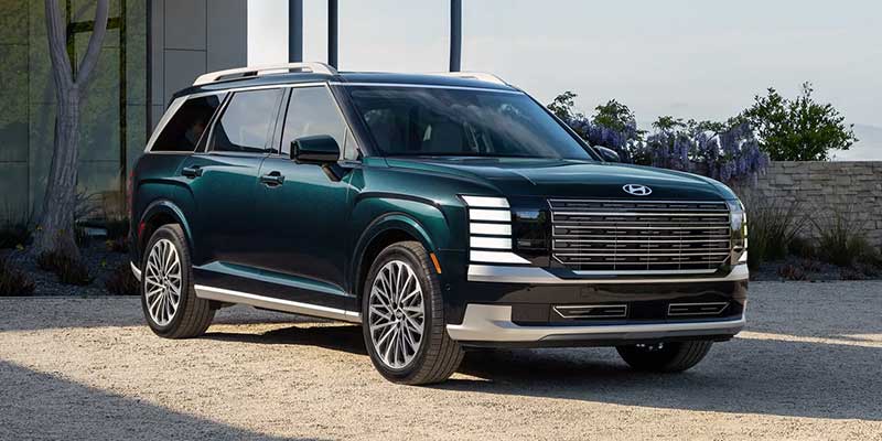 Dark green SUV with a chrome grille, light bar headlights, and roof rails, parked on gravel