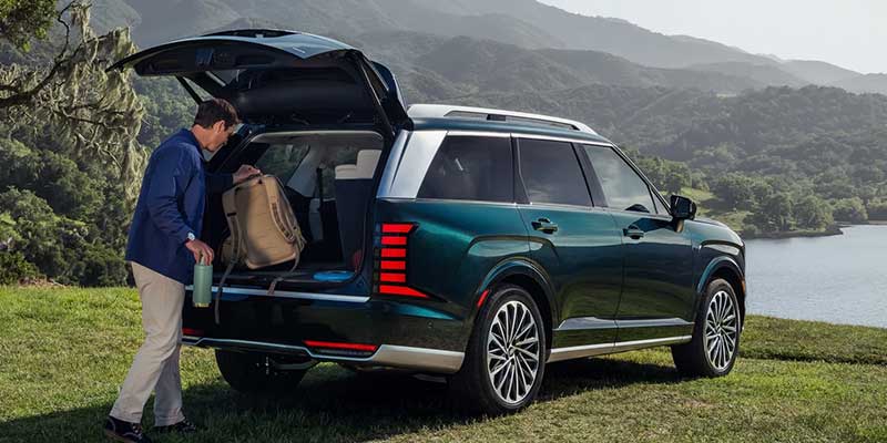 Man loading a backpack into the open trunk of a dark green SUV, overlooking a lake and mountains