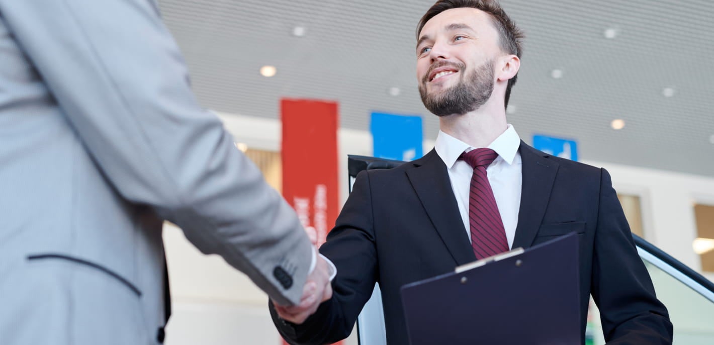 Car dealership sales representative shaking hands with a customer while holding a clipboard.