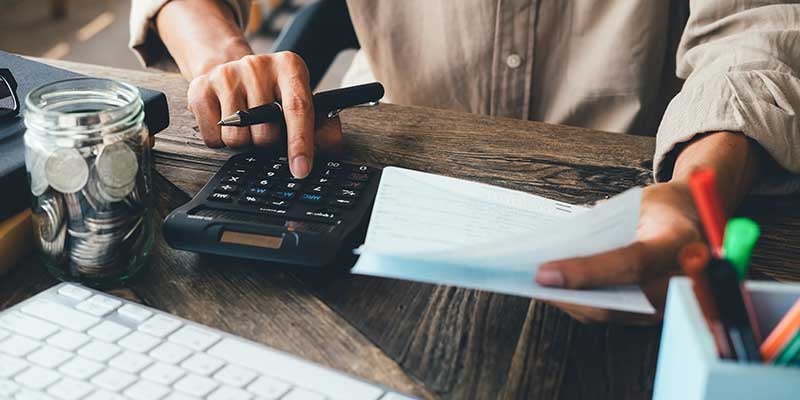Hands using a calculator to balance finances, with a jar of coins, papers, and a keyboard nearby