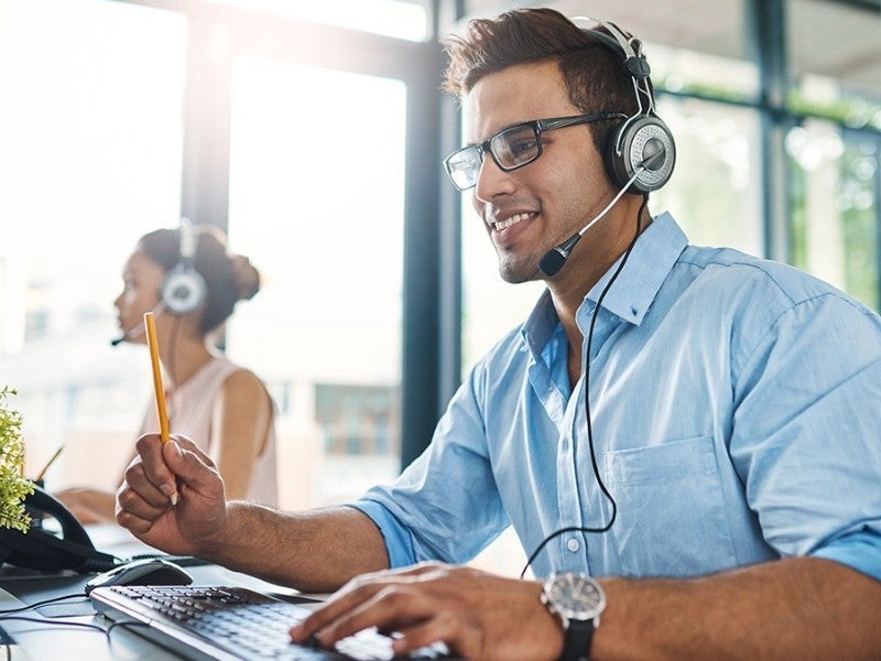 Smiling man wearing headset with microphone, glasses, and blue shirt, typing at a computer in an office