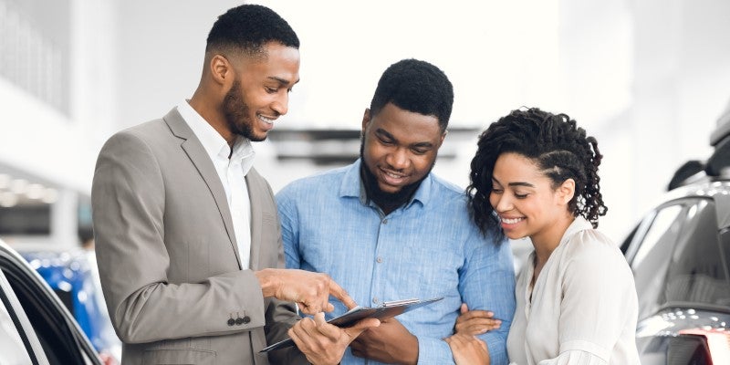 A salesperson points to a document on a clipboard for a smiling couple in a car dealership