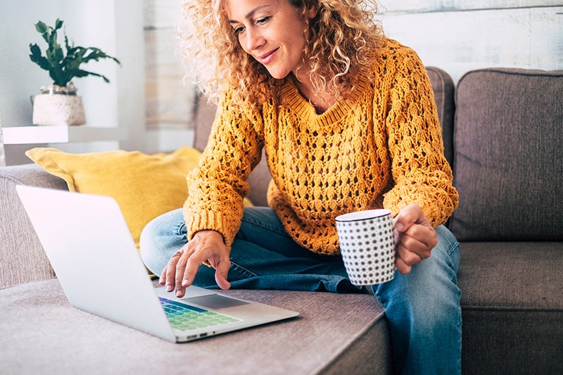 A smiling woman with curly hair, in a yellow sweater, sitting on a couch, using a laptop and holding a mug